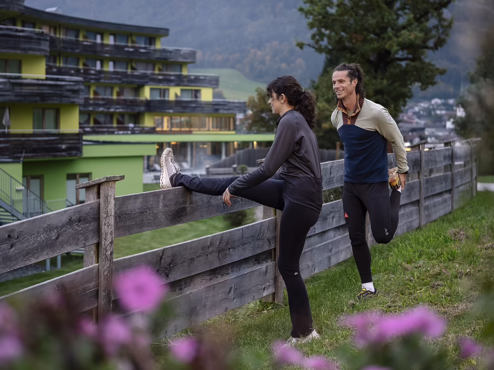 Zwei Personen dehnen sich an einem Holzzaun mit Blick auf das Hotel und die Berge