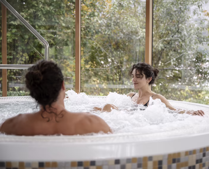 Zwei Personen entspannen im Whirlpool im Hotel DAS SIEBEN mit großen Fenstern und Blick ins Grüne