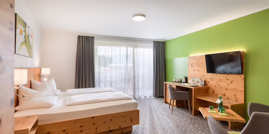 Großzügiges Hotelzimmer mit Doppelbett, grün gestrichener Wand, Schreibtisch und gemütlicher Sitzecke