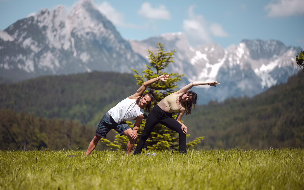 Zwei Personen üben Pilates auf einer Bergwiese vor dem Tiroler Alpenpanorama.