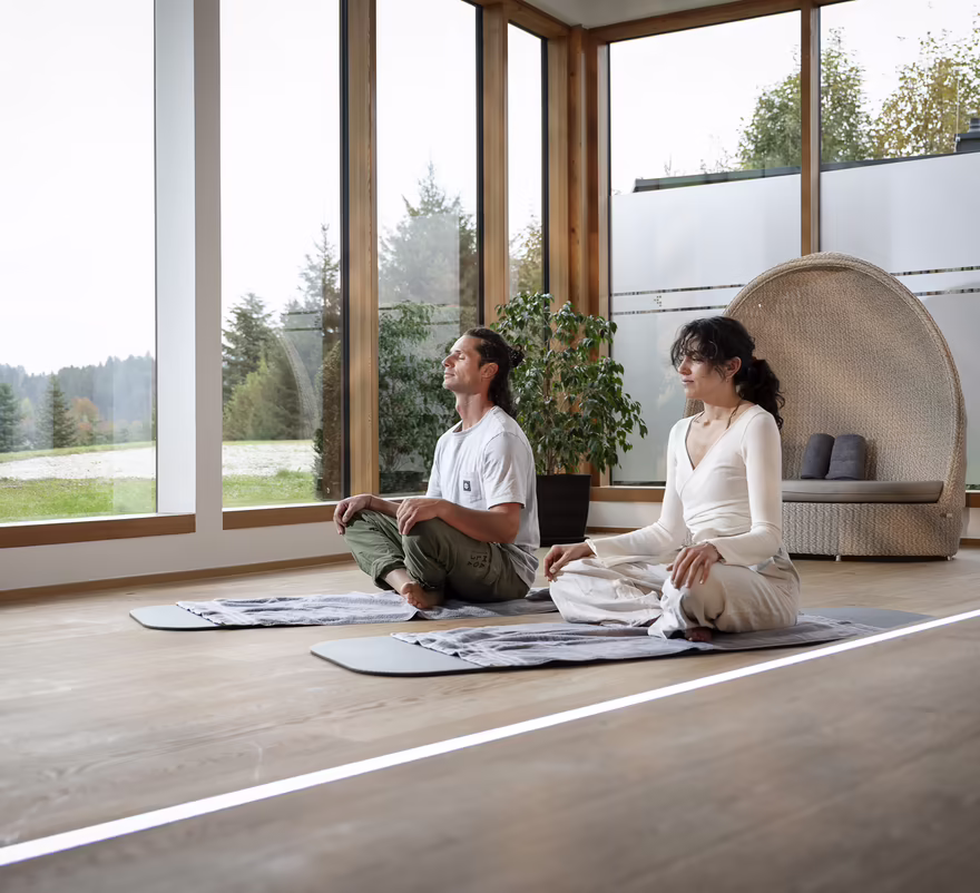 Zwei Personen sitzen auf Yogamatten in einem hellen Raum und meditieren
