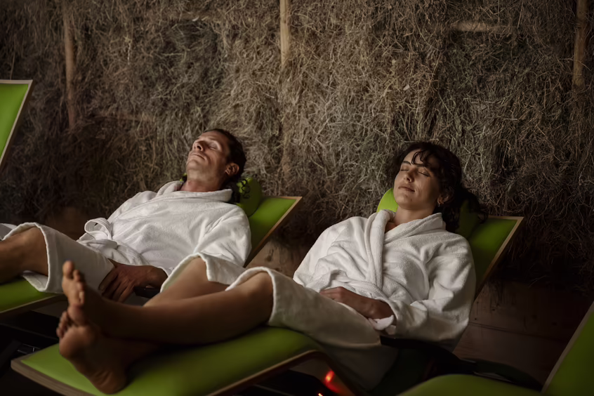 Zwei Personen liegen entspannt auf grünen Wellnessliegen vor einer Heuwand