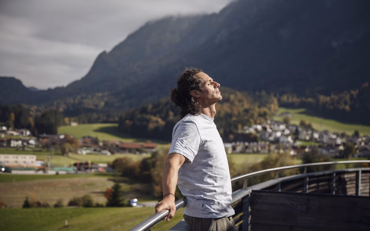 DAS SIEBEN Vital Journal | Warum Wellness mehr ist als Entspannung ©alexgretter Mann atmet auf einer Terrasse mit weitem Blick auf Berge und Landschaft im DAS SIEBEN durch